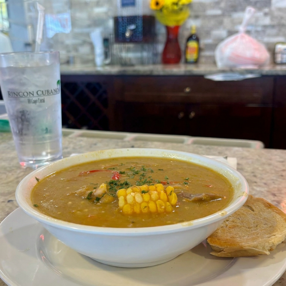 Bowl of hearty soup topped with corn and herbs, served with bread at a Cuban restaurant counter.